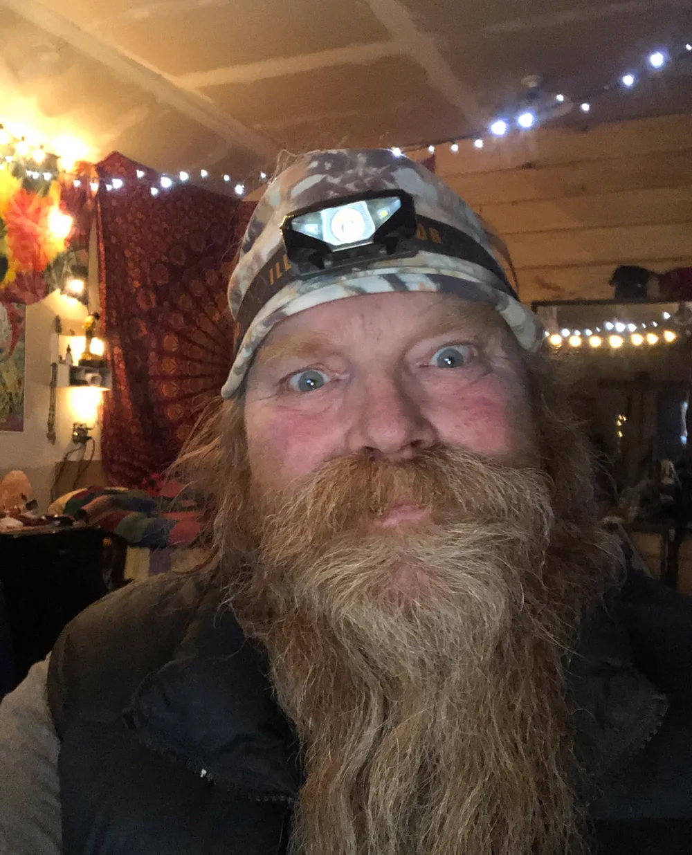 Rusty in a camo knit cap with a headlamp, a long red beard and wide eyes, string lights and tapestries behind him.